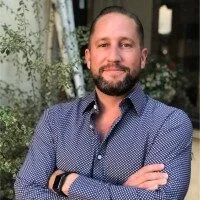 Confident man in blue patterned shirt smiles, standing in a welcoming, natural setting.